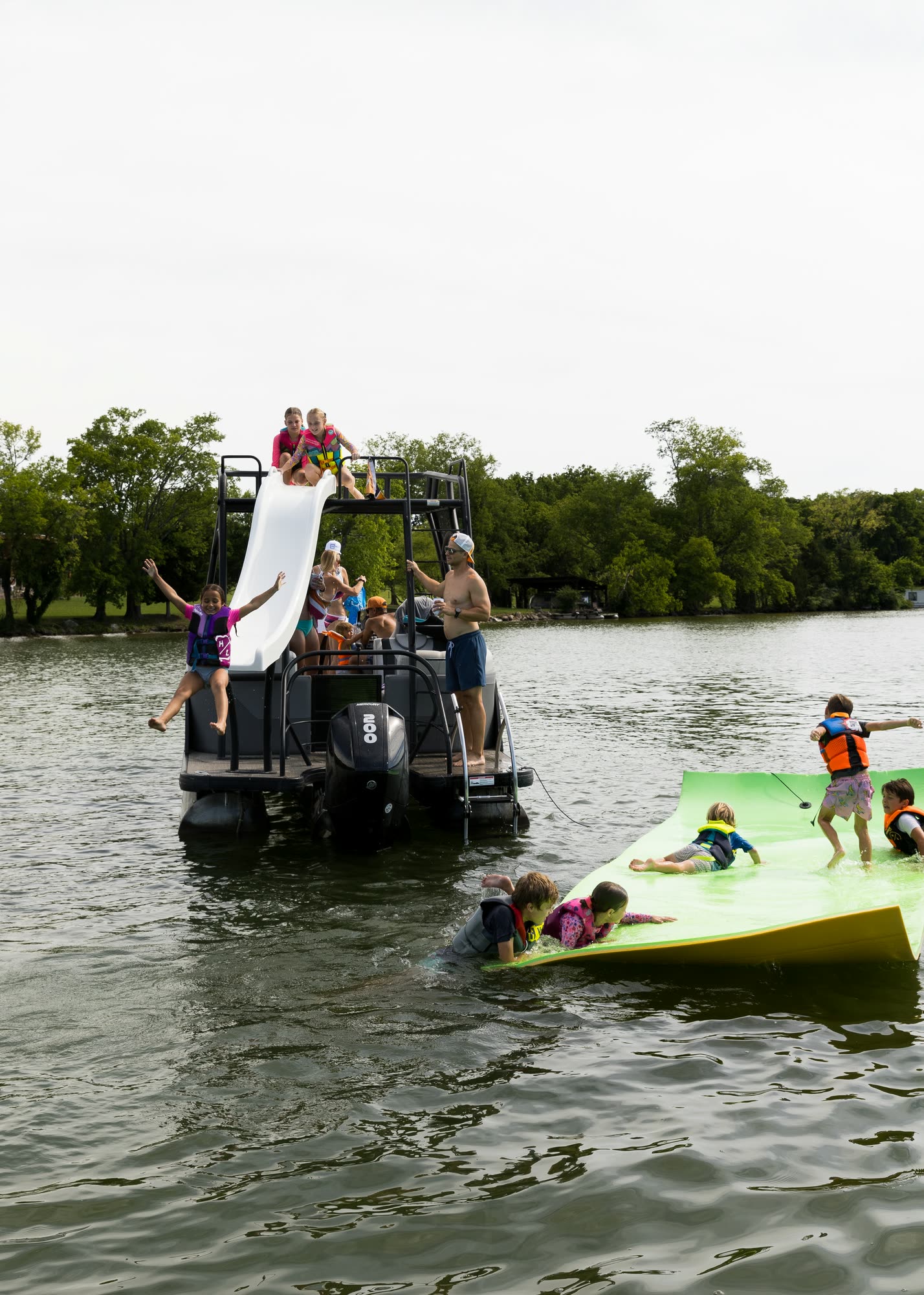 Children playing on a foam float mat and a slide beside a double-decker pontoon boat