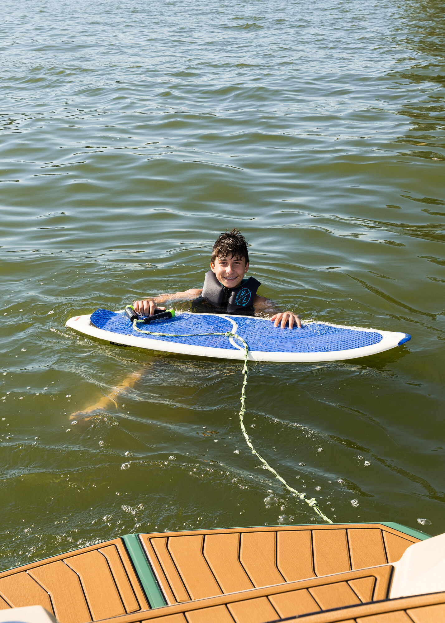 A young rider floating beside a blue wakesurf board in a life jacket at the stern of a boat