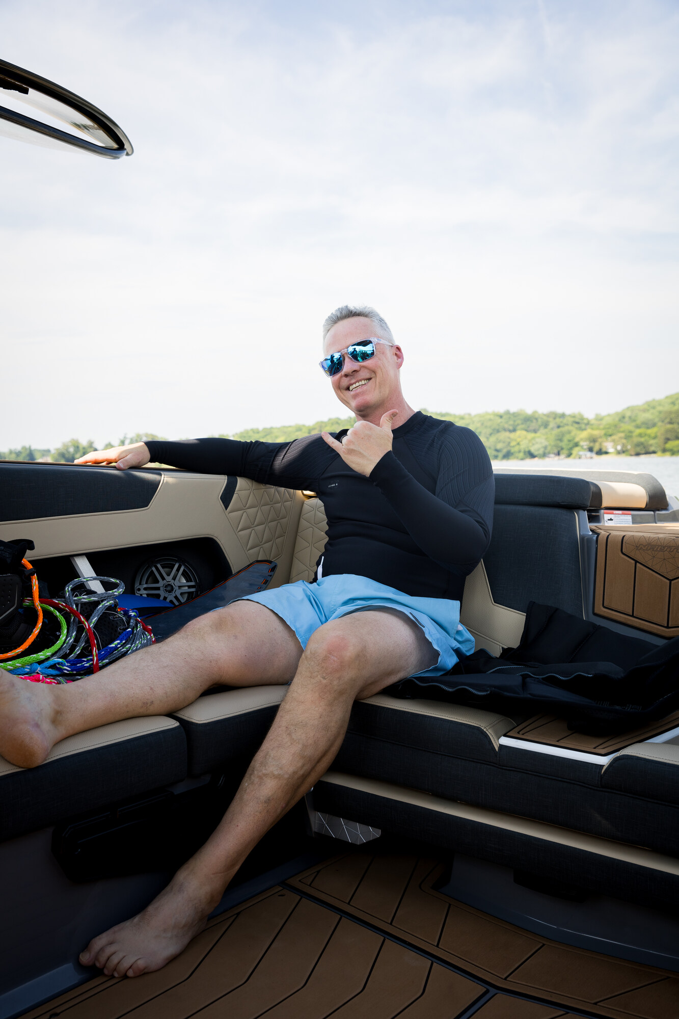 An adult in a black rash guard seated in a boat, smiling and giving a hang-loose sign